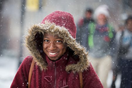 Student wearing a parka with her hood pulled up walks through the snow on front campus