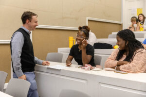 Professor Kenaston stands in front of a desk and speaks with two students.