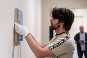 Male art history student wearing  white gloves hangs a painted canvas on the wall in a gallery at the Maier Museum of Art at Randolph College.
