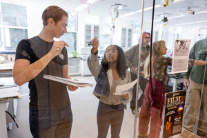 Two students stand and discuss an equation written on a glass panel with other more students in the background.