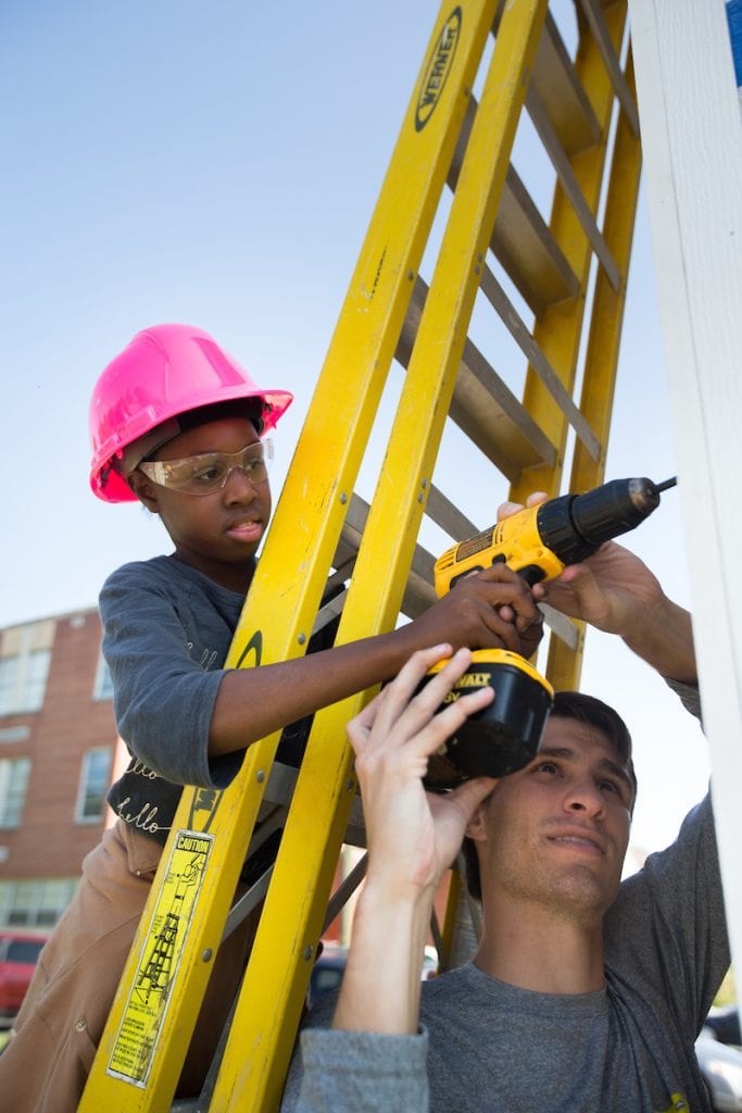 Austin Collier '19 helps a middle school student use a cordless drill while putting up walls for a tiny house.