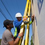 A Randolph student holds the ladder while a middle schooler drills the siding of the tiny house.