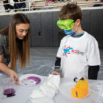 Female Randolph student teaches a science experiment to a young boy.