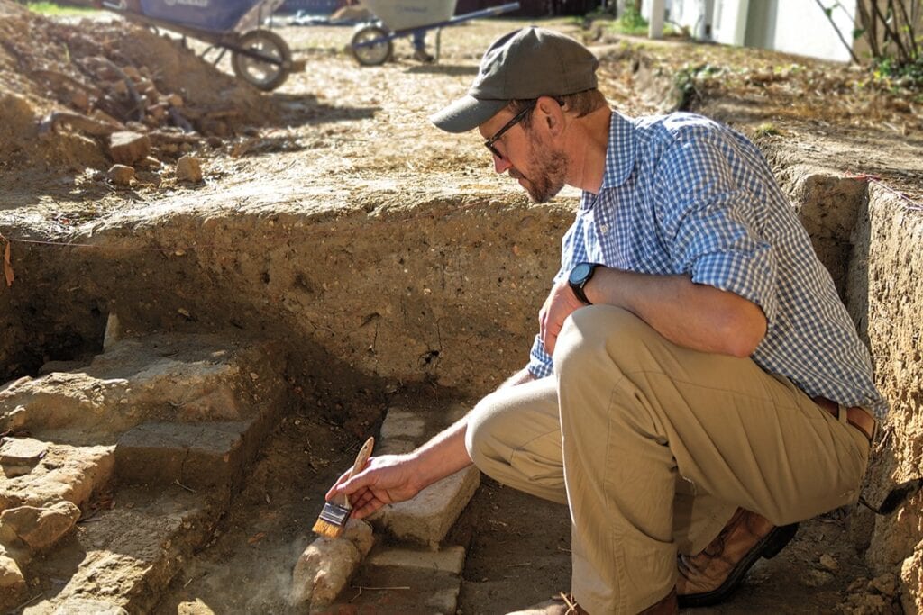 Jack Gary works at an archaeological dig, kneeling in the dirt with a brush.