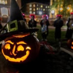 A lit-up, carved pumpkin is in the foreground of this image, with students gathered behind it, arms around each other in a circle, during Pumpkin Parade