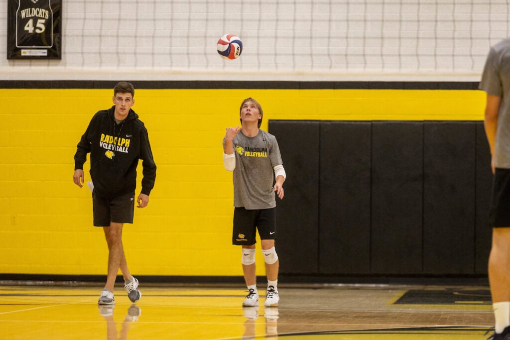 A men's volleyball player tosses the ball up in the air while coach Jordan Leandro looks on