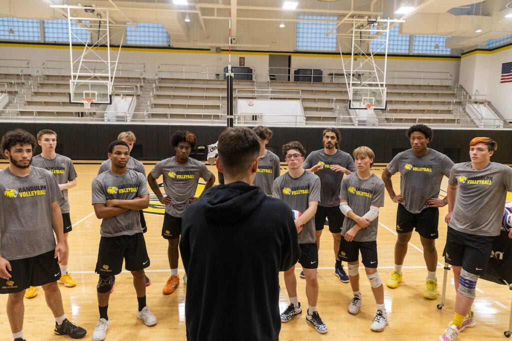 Men's volleyball coach Jordan Leandro talks to his team. His back is to the camera, while all of his players look at him. 