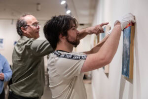 Museum preparator John Spanich works with a student hanging a piece of artwork in the Maier
