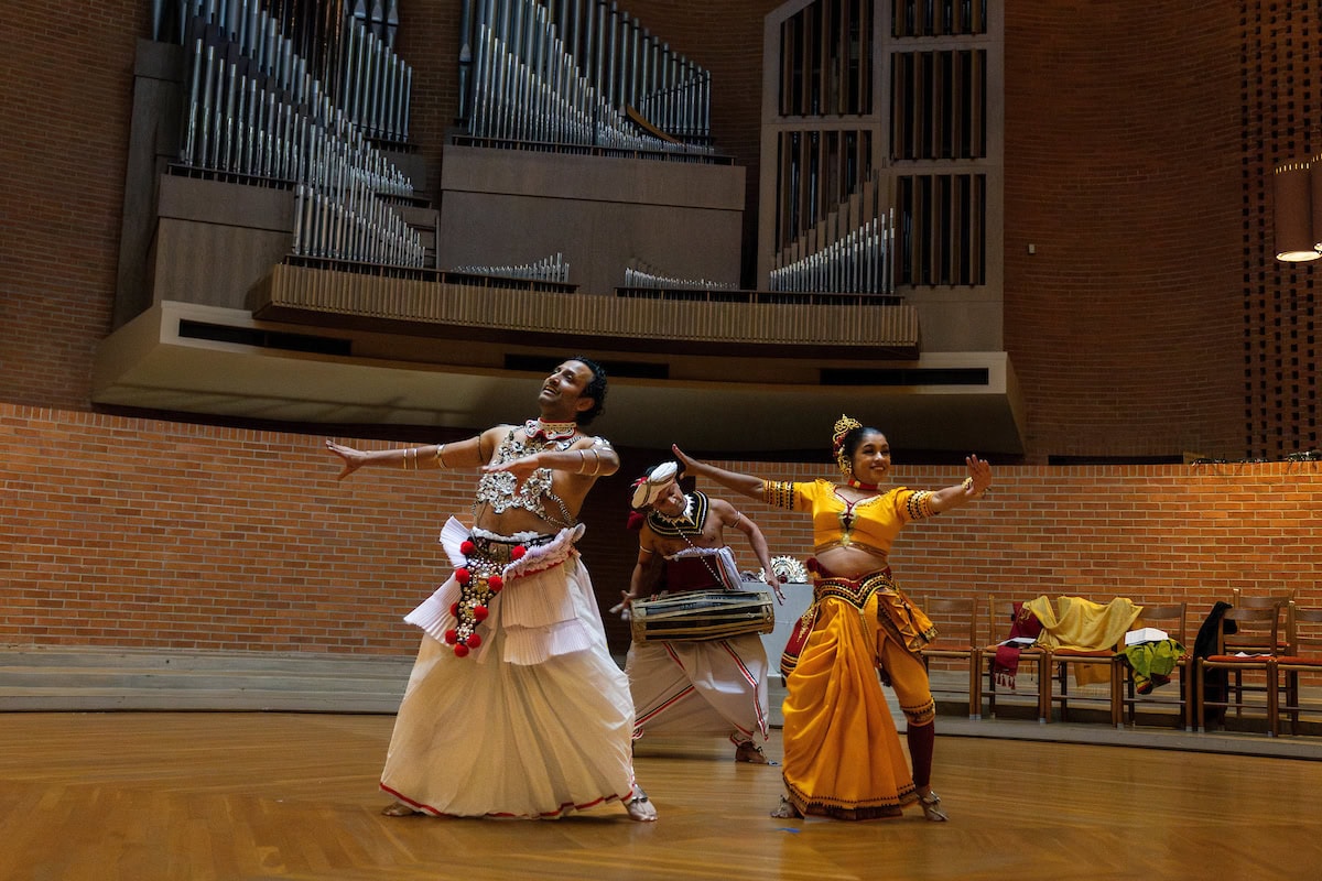 Sudesh Mantillake, Chinthaka Bandara and Vajira Mantillake perform in Houston Memorial Chapel. Sudesh wears a traditional Sri Lankan Kandy dance ensemble, with a jeweled chestplate and bangles up and down his arms. Vajira is in a bright yellow sari-like dress, and Chinthaka stands behind them with a drum. 