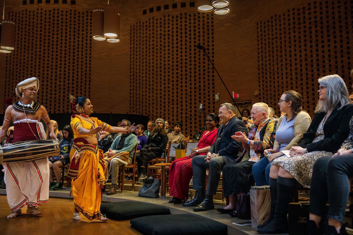 Vajira Mantillake stands in front of the crowd, wearing a yellow sari-like dress, while Chinthaka Bandara stands next to her, playing the drum around his waist