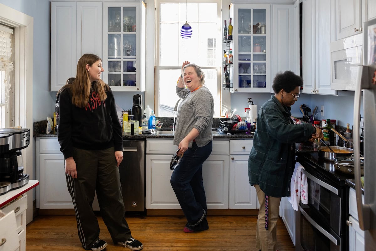 Professor Sarah Sojka stands in the middle of her kitchen, laughing, as two of her students stand nearby. One looks at her, and the other is cooking at the stovetop as part of her "adulting" class, teaching students life skills.