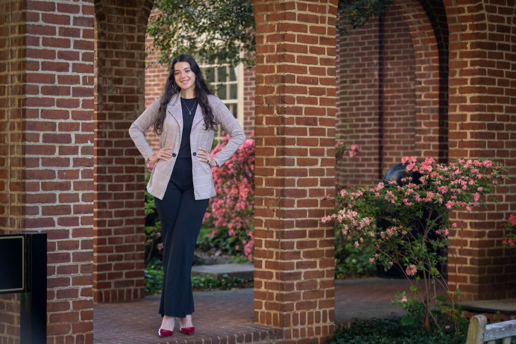 Lexie Lama poses outside on the sidewalk by the library, standing next to one of the brick pillars Lexie Lama poses outside on the sidewalk by the library, standing next to one of the brick pillars