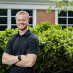 Cole Torres '25 poses for a portrait in front of the Michels Athletic Center. He's in a black collared shirt with his arms crossed. Cole Torres '25 poses for a portrait in front of the Michels Athletic Center. He's in a black collared shirt with his arms crossed.