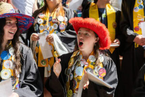 A group of students, several in their robes and button-covered stoles, sing during Daisy Chain