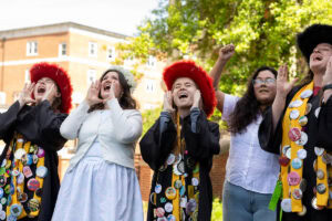 A group of students, several in their robes and button-covered stoles, sing during Daisy Chain