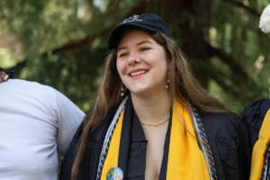 Shauna Shepard '25 smiles during the Daisy Chain ceremony, wearing her robe, yellow stole, and a hat.