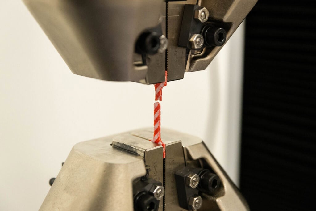 A small, 3D printed prototype, made of red and white stripes, sits broken in half in a machine 