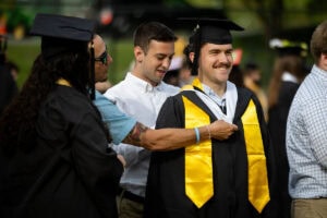 A student, wearing a cap, gown and yellow stole, smiles as his squire places his hood