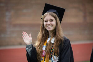 A student wearing a cap and gown smiles and waves to the camera 