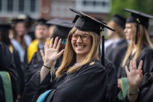 A student in a cap and gown smiles and waves to the camera 