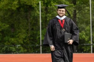 A student in a cap and gown smiles, diploma in hand, as he walks off the Commencement stage 