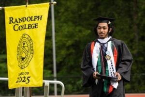 A student in a cap and gown smiles, diploma in hand, as he walks off the Commencement stage 