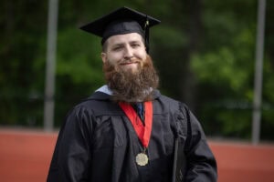 A student in a cap and gown smiles, diploma in hand, as he walks off the Commencement stage 