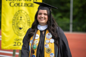 A female student in a cap, gown, and yellow stole with buttons, smiles while walking onstage during Commencement 