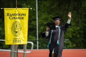 A graduate in cap and gown waves to the crowd, with a Randolph College flag in the corner of the photo 