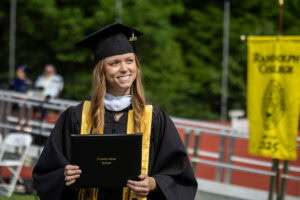 A female student in a cap, gown, and yellow stole smiles while holding her diloma 