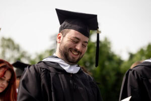 A male student in his cap and gown smiles while looking down during Commencement 