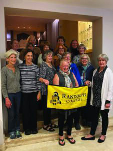 A group of women poses together, with one holding a yellow Randolph College flag in front