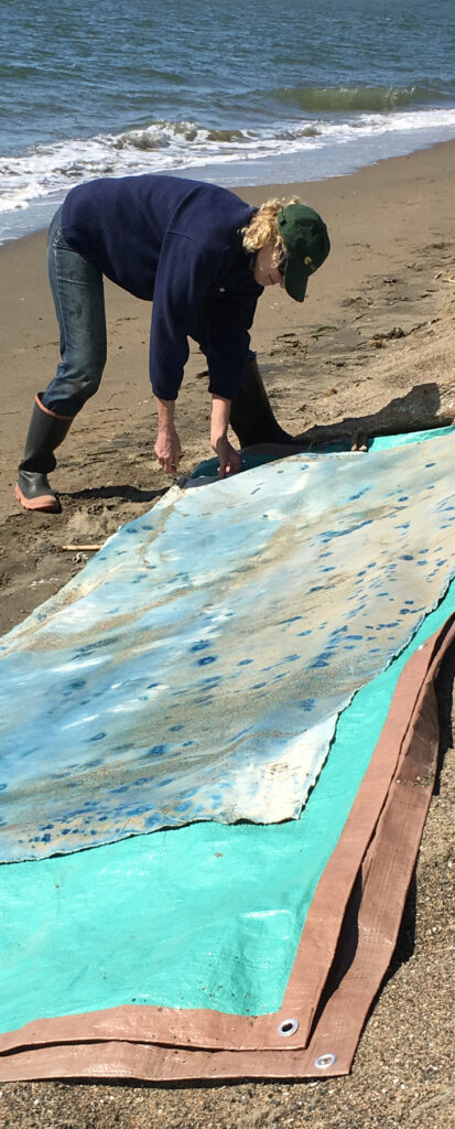 Ann Holsberry works on a piece of art on the beach. The canvas is laid out in front of her, and she leans over it.