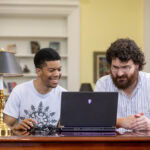 A student and a professor sit together, both looking at a laptop.