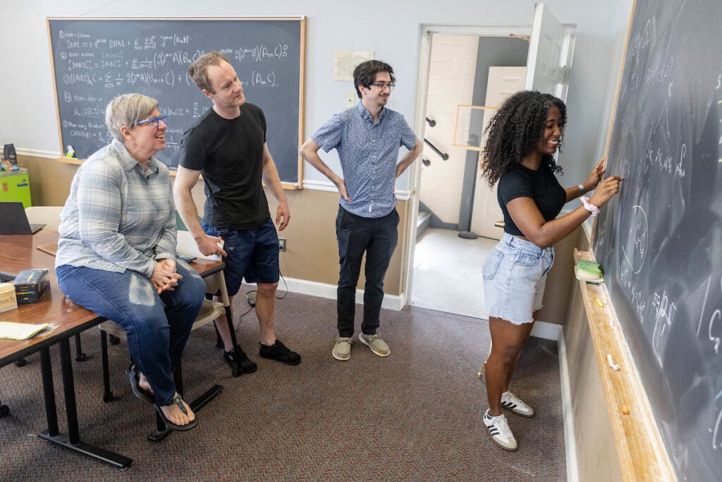 A group of people standing around a blackboard, while three people watch from behind her. A group of people standing around a blackboard, while three people watch from behind her.
