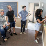 A group of people standing around a blackboard, while three people watch from behind her.