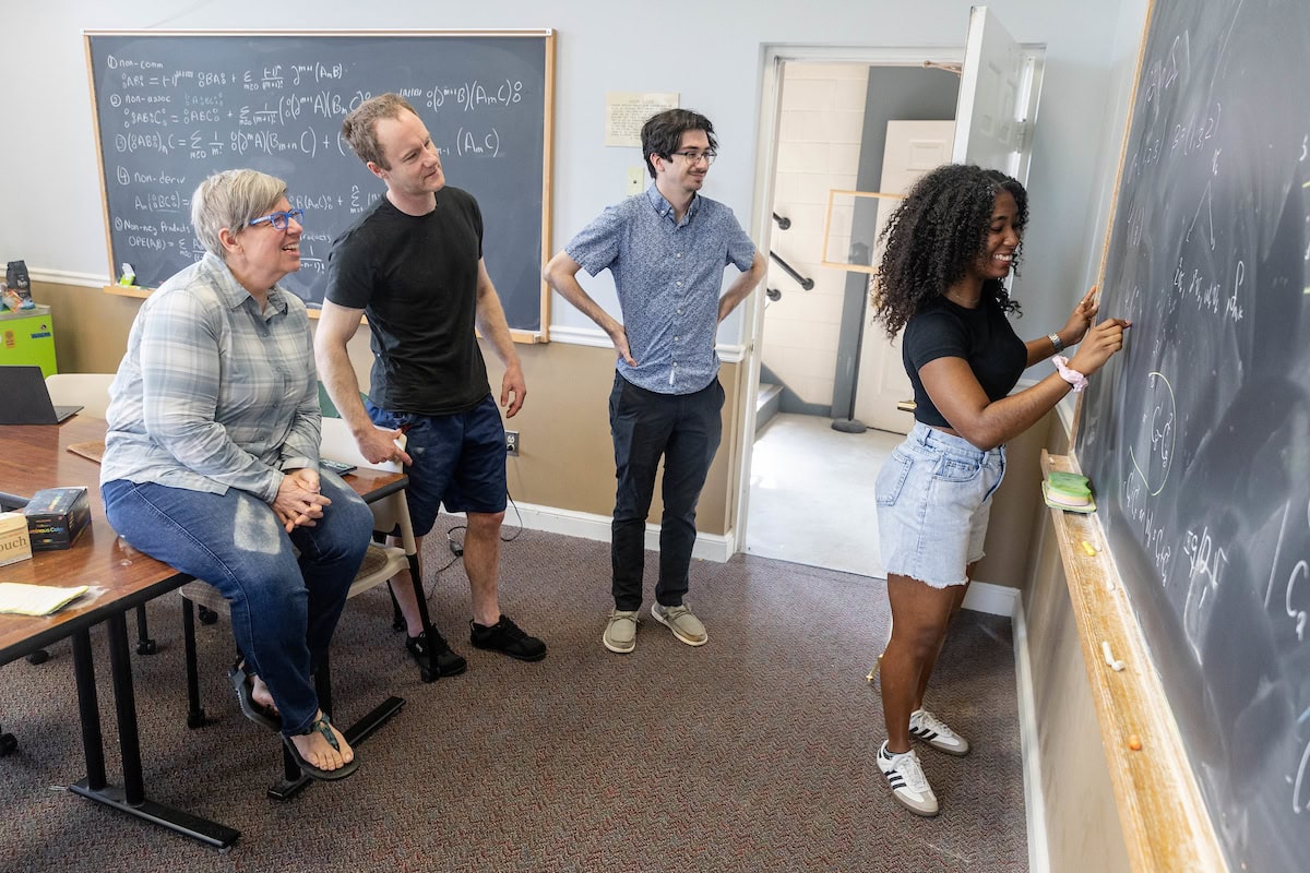A group of people standing around a blackboard, while three people watch from behind her.