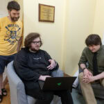 Three men--two sitting and one standing--talk, with one holding a laptop Three men--two sitting and one standing--talk, with one holding a laptop