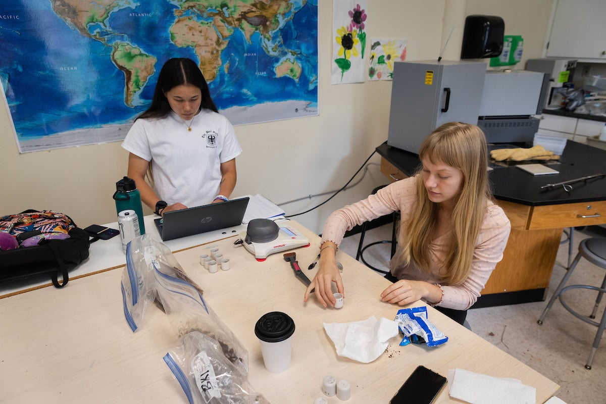 Two women work in a lab together, one on a laptop and the other working directly with samples. 