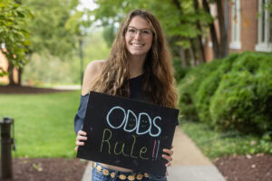 Student poses with a chalkboard that says "Odds Rule" 