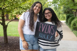 Two students pose with a chalkboard that says "Evens Rule"