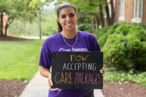 A student poses with a chalkboard that says "Now accepting care packages" 