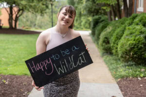 A student poses with a chalkboard that says "Happy to be a WildCat"