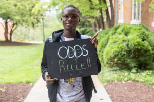 A student poses with a chalkboard that says "Odds Rule" 
