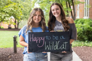 Two students pose with a chalkboard that says "Happy to be a WildCat"