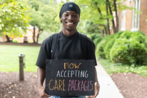 A student poses with a chalkboard that says "Now accepting care packages"