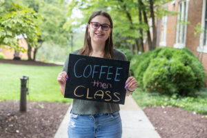 A student poses with a chalkboard that says "Coffee then class"