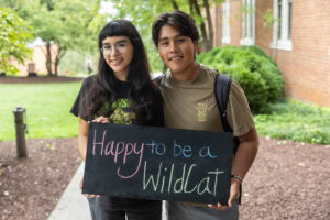 Two students pose with a chalkboard that says "Happy to be a WildCat"