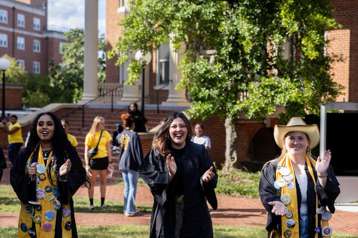 Three students walk on campus clapping their hands 
