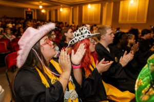 Students in their graduation gown and stoles, with crazy hats on, clap in the audience 